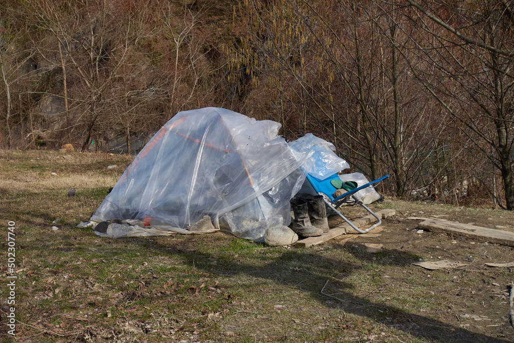 Parking of the homeless on the bank of a mountain river. The tent is covered with plastic wrap. A folding chair and boots stand in the mud at the entrance.
