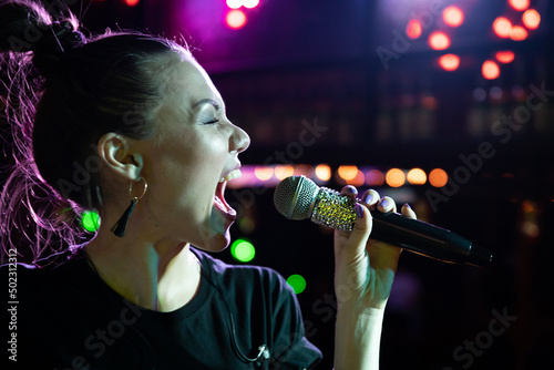 Young singing woman with microphone having fun on open stage in karaoke club.