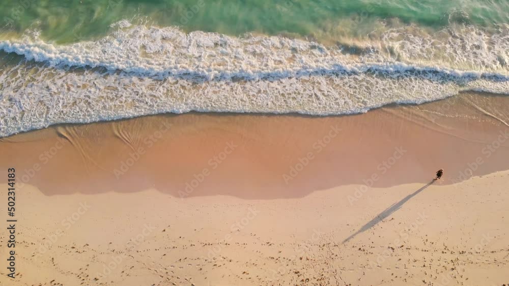 Overhead view of alone man walking by the white sandy beach along the amazing clear light blue ocean. Aerial view of a walking man on a beach, lonely on the sand with beautiful view of waves.