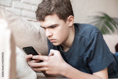 A teenage boy lies on the couch with a smartphone in his hands. A young man with eyes widened with horror or surprise. Emotions.