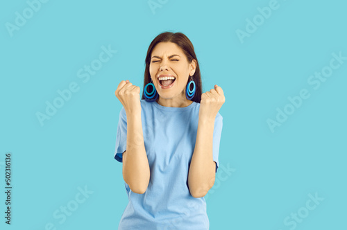 Beautiful young woman wins cool prize or is excited about holiday sale. Happy euphoric lady in blue T shirt and earrings standing on blue background, fist pumping and screaming YES, I DID IT, HOORAY