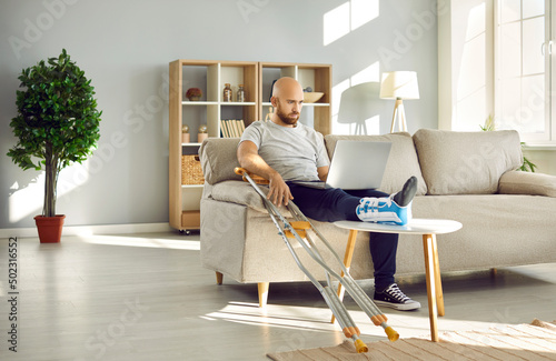 Fotobehang Sick businessman with broken leg in bandage sit on sofa at home work on laptop online