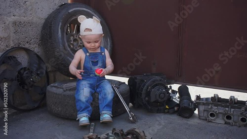 serious baby sits on tire, and around auto parts