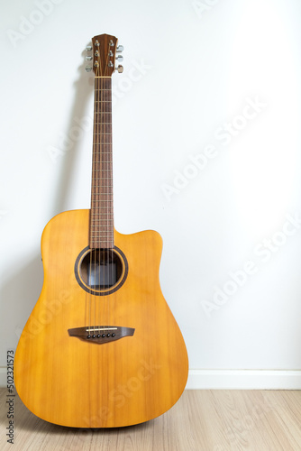 An acoustic dreadnought guitar leaned against the white wall background. Veritcal image copy space.
