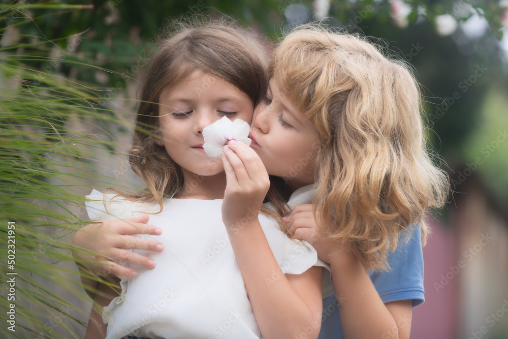 Two little children hug and kiss each other in summer garden. Kids couple in love. Romantic boy and shy girl, giving a flower for love and romance. Two little children hug and kiss each other in summer garden. Kids couple in love. Romantic boy and shy girl, giving a flower for love and romance.