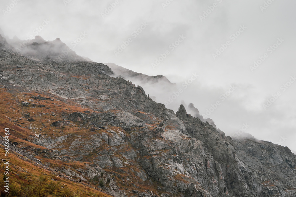 Atmospheric misty landscape with fuzzy silhouettes of sharp rocks in low clouds during rain. Dramatic view to large mountains blurred in rain haze in gray low clouds.