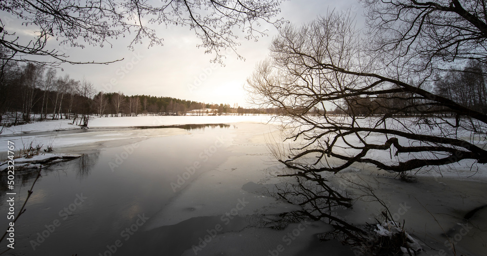 Epic spring landscape by the river. March evening, snow melts. Winding river with snow-covered banks and ice. Trees are reflected in the water.