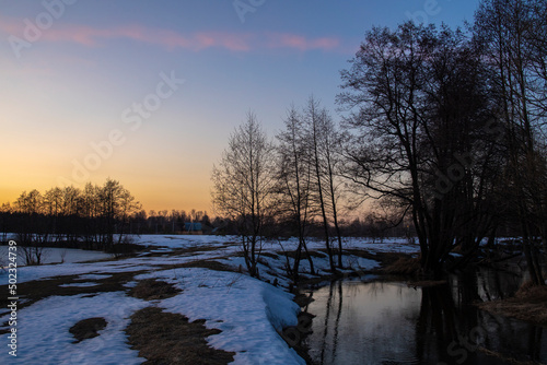 Evening spring landscape, trees by the river, snow and thawed patches. Charming landscape in early spring against the backdrop of sunset. Melting ice and snow.