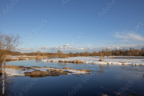 Fototapeta Naklejka Na Ścianę i Meble -  A picturesque landscape, early spring, a river with snow-covered banks, dry grass and bushes. March sunny day by the river. The first thaws, the snow is melting.