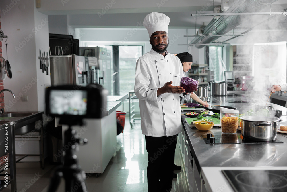 African american head chef having red cabbage filming cooking of gourmet dish while looking at camera. Culinary vloggers in restaurant kitchen recording culinary course for internet and TV broadcast.