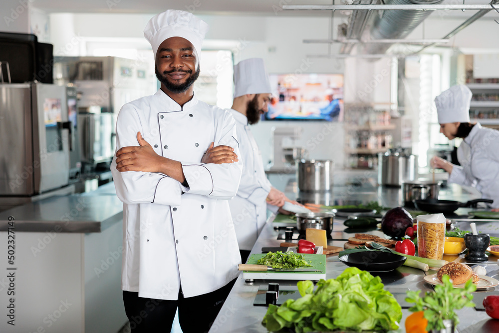 Gastronomy expert standing in restaurant professional kitchen with arms crossed while smiling at camera. Confident head chef wearing cooking uniform while preparing ingredients for dinner service.