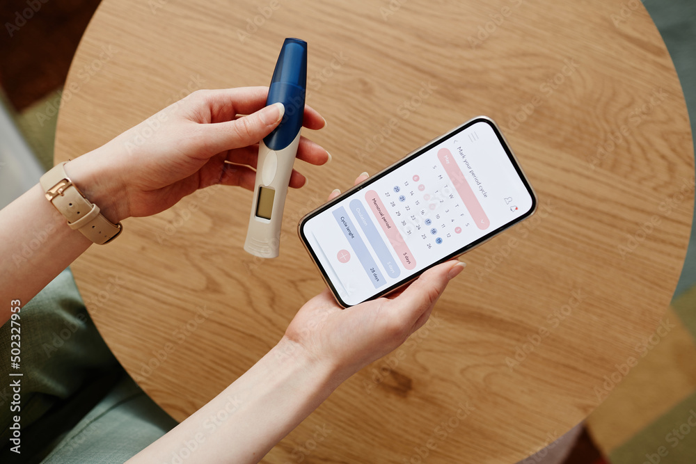 Close up of young woman calculating menstrual cycle using mobile app and holding pregnancy test - obrazy, fototapety, plakaty 