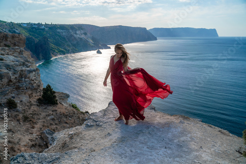 A girl with flowing hair in a long red dress stands on a rock above the sea. The stone can be seen in the sea. Sunny path to the sea from the sun.