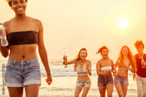 african american woman holding plastic bottle of water   behind her group of mixed race internation friends dancing and having fun with lemonade on sunset beach