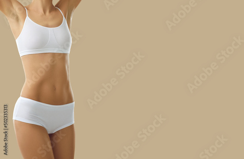 Slender woman with fit body posing in white base lingerie collection isolated on beige background. Cropped view of unknown woman standing with raised hands in top and panties near copy space. Banner.