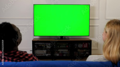Green screen TV set laughing multiethnic family couple looking at TV chroma key mock up display. Back view husband and wife watching comedy movie series on TV. Man and woman sitting on couch at home
