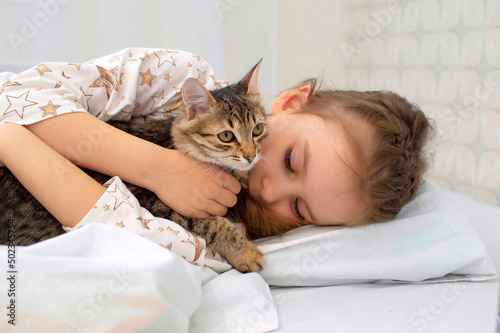 Portrait. Close-up. The child is resting on the bed with his kitten. A little girl in pajamas on a sunny morning with tenderness and love hugs a gray cat and smiles