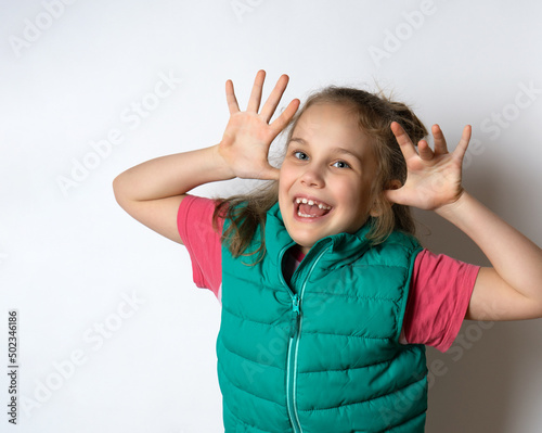 Photo portrait of a little playful child girl in a warm vest and t-shirt, grimacing, fooling around, showing her tongue on a light background.