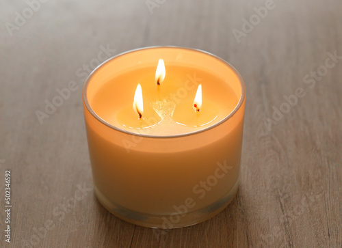 Photography A large scented candle with three burning wicks and melting wax in a glass standing on a wooden table