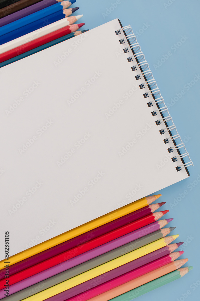 Notepad with a white blank sheet surrounded by colored pencils on a blue background. School. Stationery store. For drawing. View from above. Layout of stationery. Place for text.