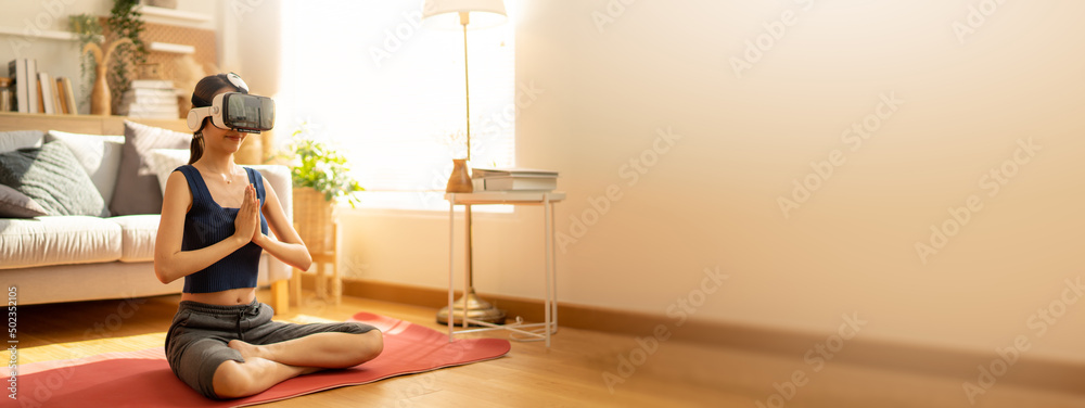 Asian Female woman wearing virtual reality glasses meditating in lotus position at home,yoga namaste enjoying simulation and relax in metaverse world.AR,VR Virtual Future technology concept
