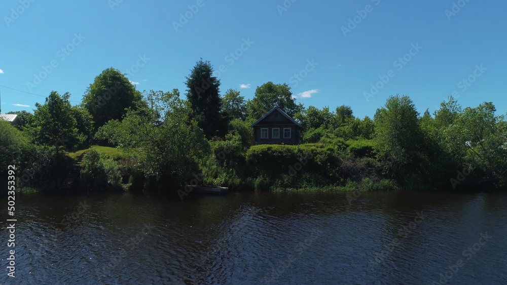 Old wooden house located on the shore of a river in the countryside. Shot. Small calm river among the forest and a village house on blue clear sky background.