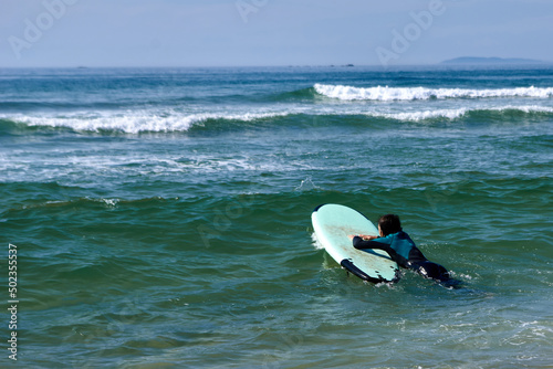 Young surfer in a black and green wetsuit is lying on a surfboard to ride the waves of a beach and surfing photo