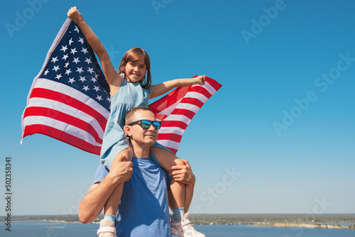 Happy family father and daughter sitting on father's shoulders with America flag enjoy nature. Freedom concept