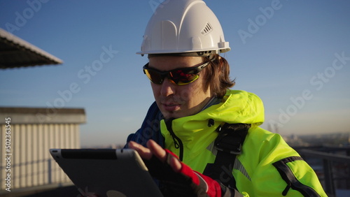 Worker in white helmet with rope across shoulder stands on roof. Industrial climber collects information and enters data into tablet against city closeup © wifesun