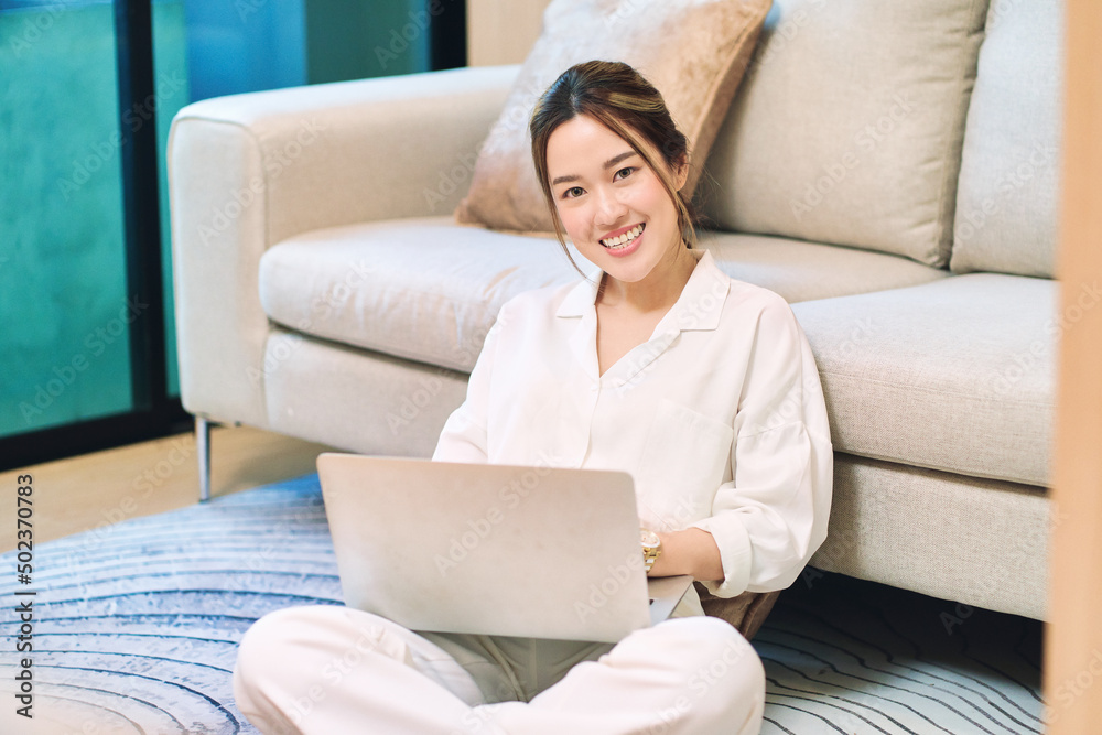 Attractive young asian woman using laptop computer while sitting near a couch at home office Attractive young asian woman using laptop computer while sitting near a couch at home office