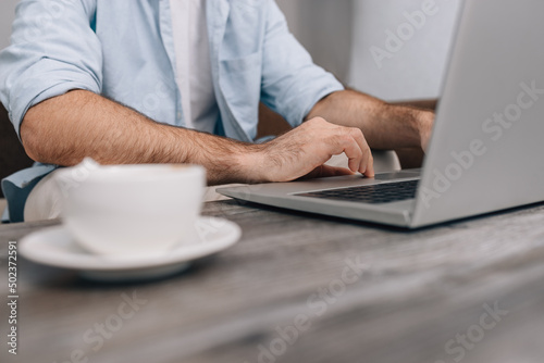 Hands of modern programmer or office manager over laptop keyboard under work on business plan. A guy in a blue shirt and white T-shirt. Cup of coffee on the table. Freelance