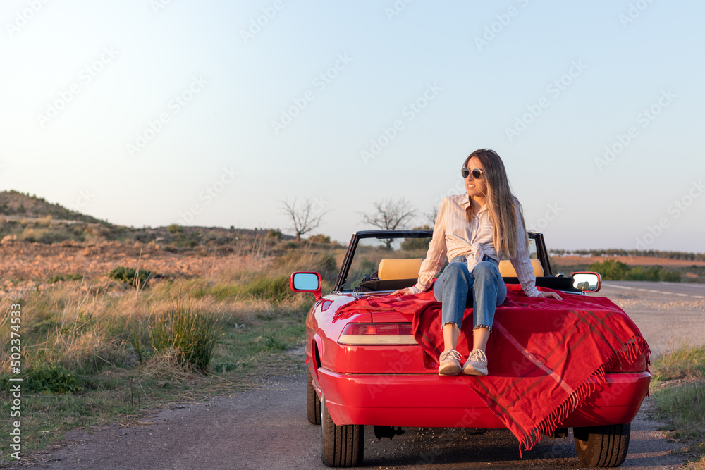 Young beautiful girl sitting on a red classic convertible car enjoying the sunset on a remote road in the countryside