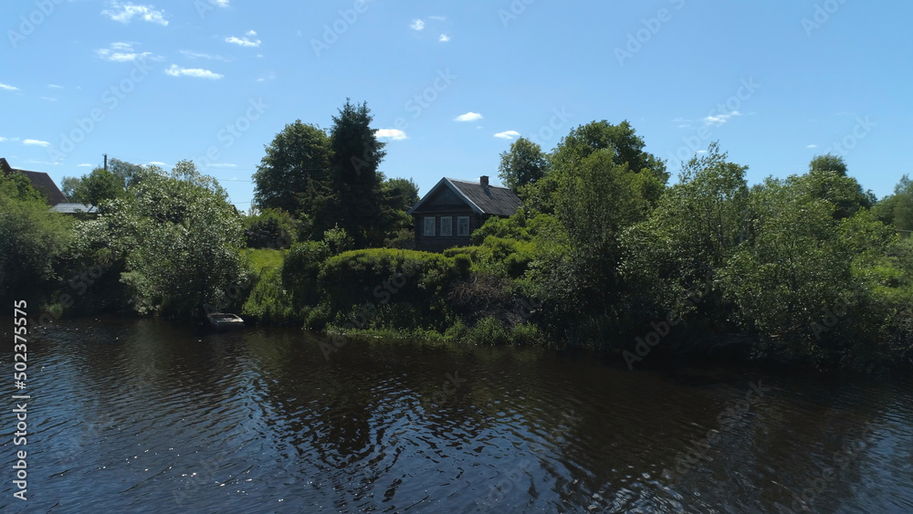 Aerial view of a river in summer village and an old wooden house. Shot. Forest and small house surrounded by green trees and bushes along the river bank on blue clear sky background.