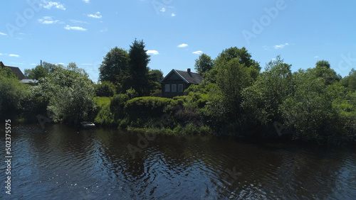 Aerial view of a river in summer village and an old wooden house. Shot. Forest and small house surrounded by green trees and bushes along the river bank on blue clear sky background.