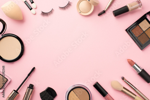 Top view photo of eyeshadow palette lipstick compact powder blush false eyelashes makeup brushes mascara lip gloss beauty blender nail polish on pastel pink background with blank space in the middle