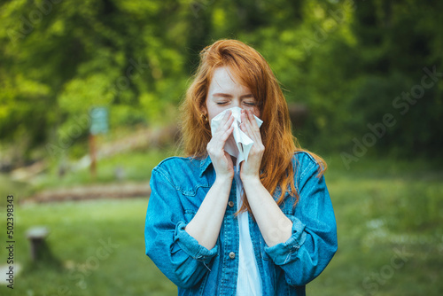 Young pretty woman blowing nose in front of blooming tree. Spring allergy concept. Attractive young adult woman coughing and sneezing outdoors. Sick people allergy or virus influenca concept.