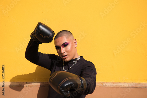 Картината върху платно Young non-binary person wears boxing gloves on a mustard yellow background, the person is make up and does different boxing poses