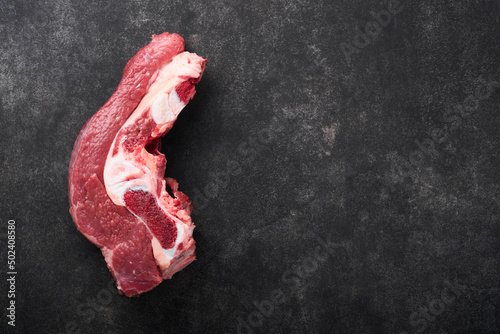 Raw beef meat. Rough piece of meat on bone for roast or soup with salt  pepper  thyme and rosemary on a black concrete background. Entrecote. Raw cowboy steak. Top view.