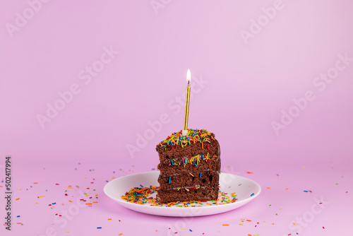 Slice of birthday cake with candle and colorful sprinkles on pink background. Chocolate cake slice, Birthday party minimal concept.