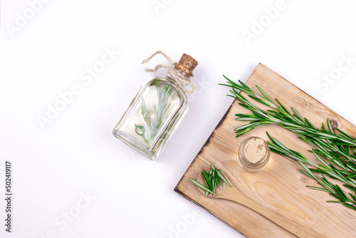 Aromatherapy rosemary oil. Wooden board, Bottle glass with rosemary essential oil, little spoon and fresh rosemary twig on white background.