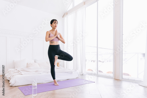 Beautiful happy young Asian woman standing in Vrksasana Posture, doing yoga exercise on purple mat with water bottle, working out in bedroom, indoor workout, Tree Pose, full length.