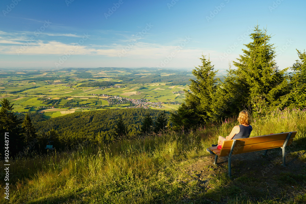 A woman sitting on a bench, looking from mount Hohenbogen to Neukirchen Heiligblut, a small town in the Bavarian Forest. A woman sitting on a bench, looking from mount Hohenbogen to Neukirchen Heiligblut, a small town in the Bavarian Forest.