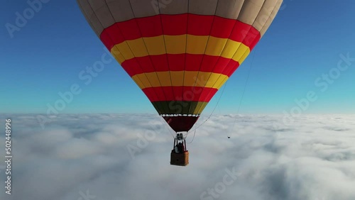 Closeup colorful red yellow hot air balloon with happy tourists flying above white clouds. First person adventure view from drone.