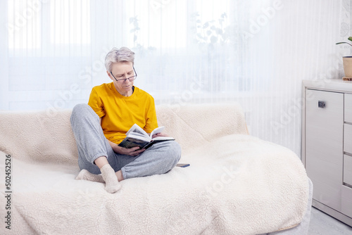 adult gray-haired woman is sitting at home on the couch and reading a book © Irina