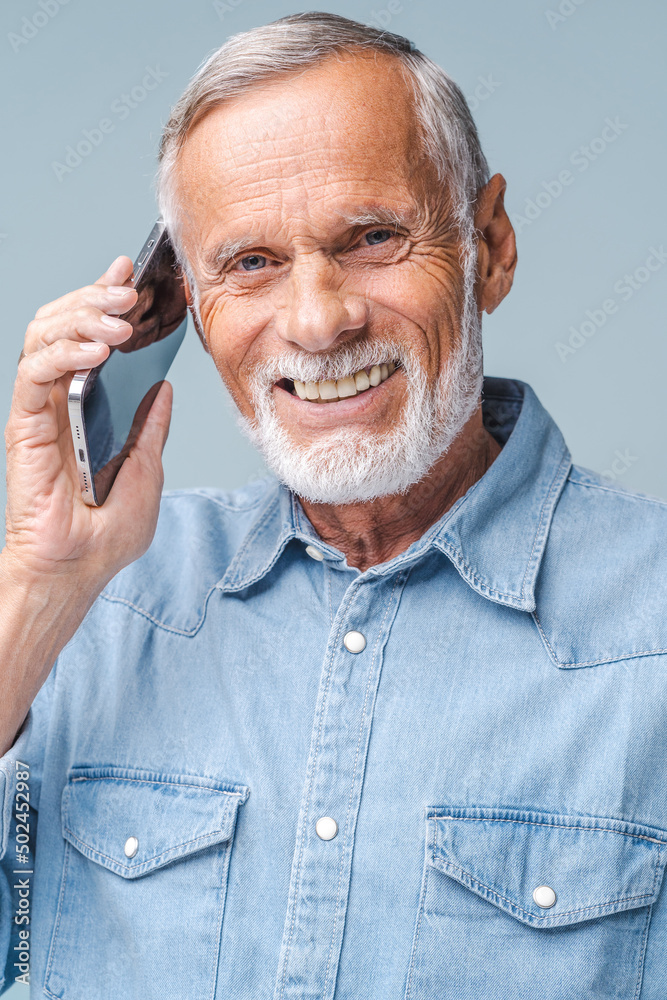 Elderly man talking phone, calling to somebody, looking at the camera with happy facial expression. Indoor studio shot isolated on blue background