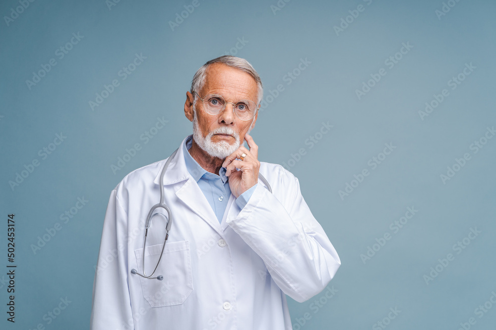 Thinking and serious caucasian senior doctor pondering about something isolated on blue background. Healthcare and medicine concept Thinking and serious caucasian senior doctor pondering about something isolated on blue background. Healthcare and medicine concept