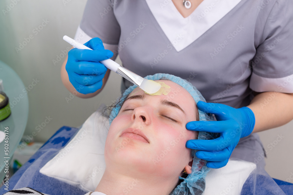 Cosmetologist applies alginate mask with spatula on face of woman.
