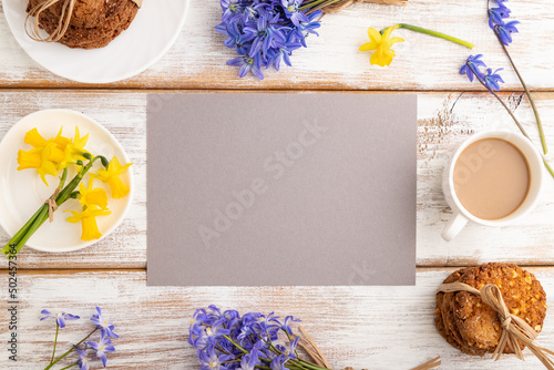 Gray paper sheet with oatmeal cookies, spring snowdrop flowers bluebells, narcissus and cup of coffee on white wooden background. top view, copy space. photo