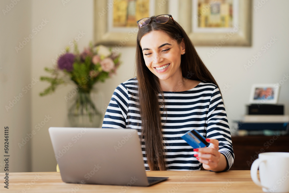 Happy woman using bank card and laptop while sitting at desk at home