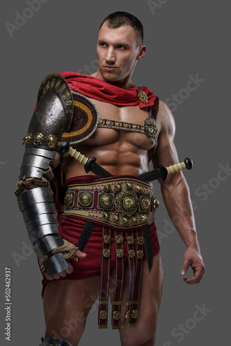Studio shot of roman gladiator with muscular build and naked torso dressed in armor and cloak.